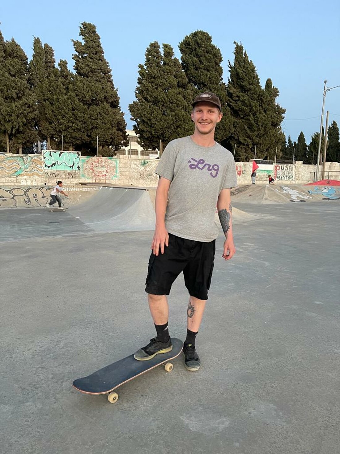 Man sieht Jörg in der Bildmitte mit dem rechten Fuß auf seinem Skateboard in die Kamera grinsen.