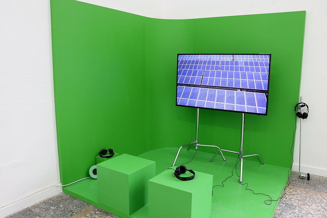 Man sieht die Ecke eines Raumes welche mit einem grünen Stoff übergezogen ist. Darauf steht ein Bildschirm welcher gerade das Bild einer Solaranlage zeigt.