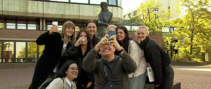 Student: innen stehen vor der Heinrich Heine Statue an der HHU und machen ein Selfie. Dabei halten einzelne von Ihnen einen Becher in der Hand, um auf Heine anzustoßen. 