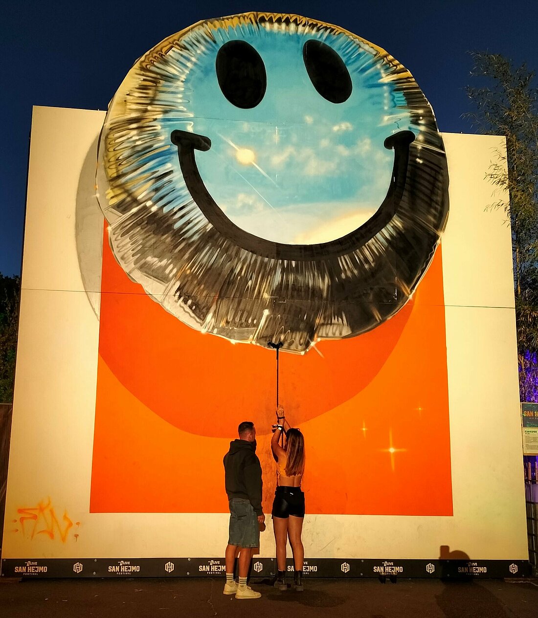 Auf dem Foto ist eine große Leinwand zu sehen. Da drauf wurde ein silberner Luftballon mit einem Smiley gemalt. Unter dem Ballon ist ein Seil gespannt. Eine Person versucht sich gerade da dran zu hängen.