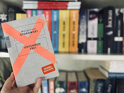 Margarete Stokowskis Buch „Untenrum Frei“ wird vor einem Bücherregal in die Luft gehalten. Das Cover des Buches ist grau mit einem orangenem Kreuz.