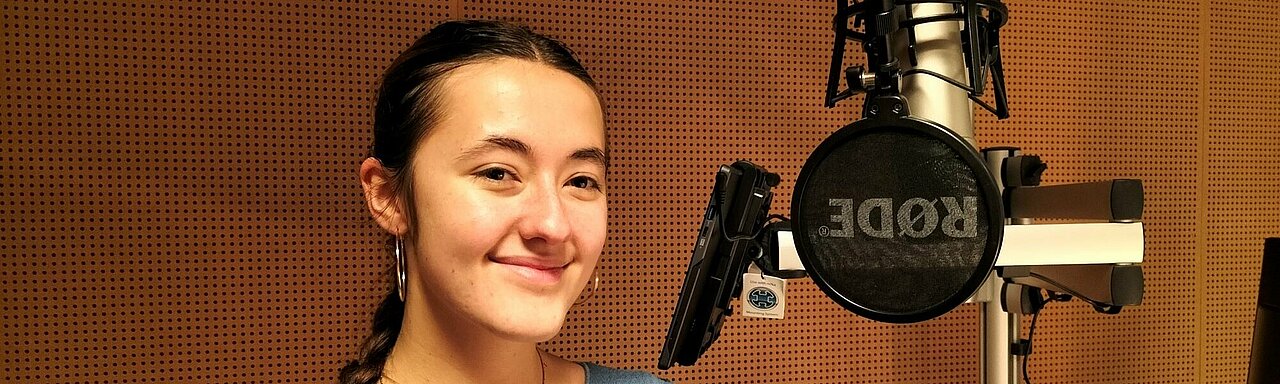 Yamina Probst steht im Studio von hochschulradio düsseldorf. Vor ihr ist ein Mikrofon zu sehen. Sie steht vor einer braunen Schallschutzwand.