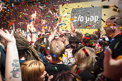 Ein Publikum feiert mit Konfetti vor dem c/o pop Festivallogo.