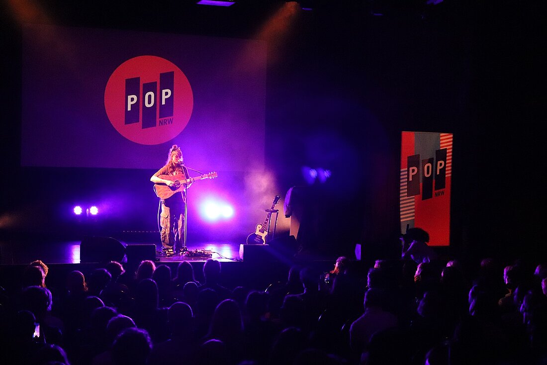 Zu sehen ist die Sängerin Philine Sonny während ihres Auftritts. Sie spielt Gitarre auf einer dunklen Bühne mit dezentem lila Licht im Hintergrund. Im Vordergrund sitzt das Publikum.