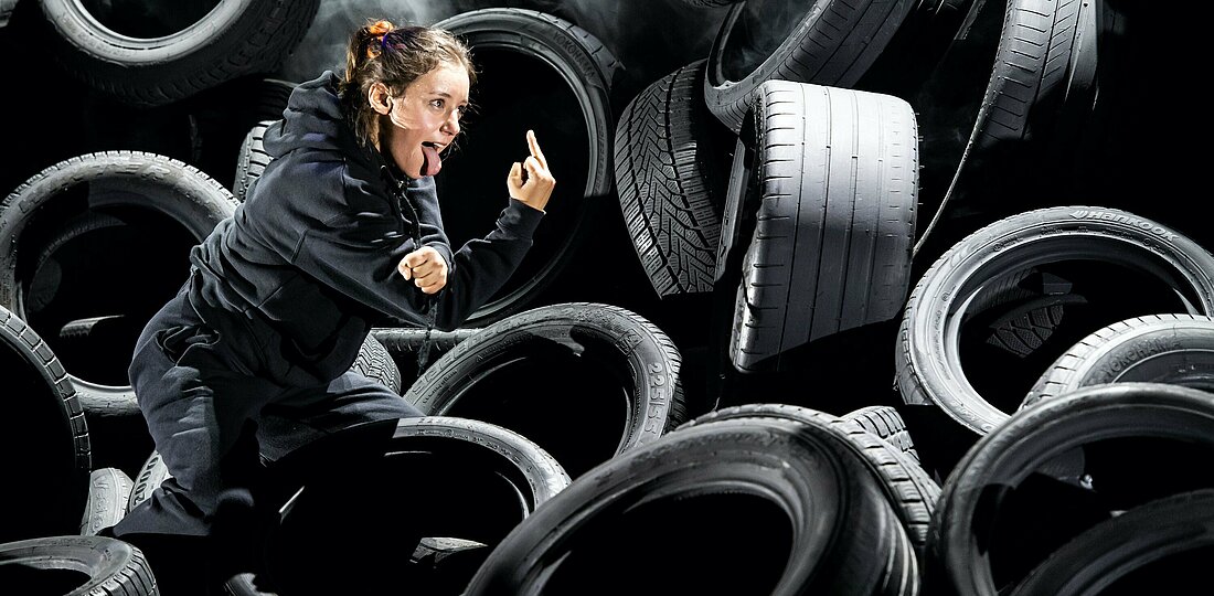 Cennet Rüya Voß kämpft gegen das E-Auto-Werk (Foto: Sandra Then). Zu sehen ist Cennet Rüya Voß auf einem Berg aus Autoreifen, wie sie die Zunge rausstreckt und den Mittelfinger zeigt.