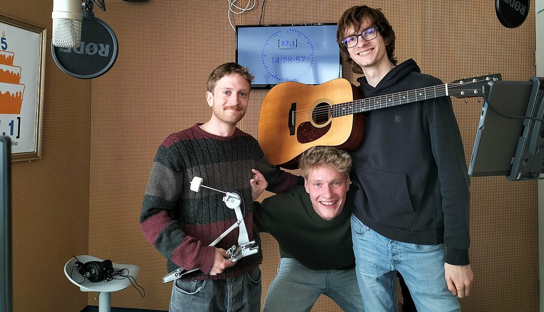 Auf dem sind von links nach rechts Carlo, Konstantin und Malte zu sehen. Carlo hält ein Drum Pedal in der Hand. Malte stützt seine Gitarre auf dem Kopf von Konstantin ab.