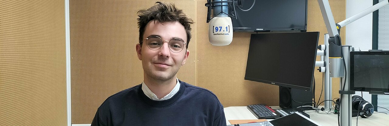 Auf dem Foto ist Josua Wienen zu sehen. Er steht im Studio, neben einem Mikrofon und lächelt in die Kamera.