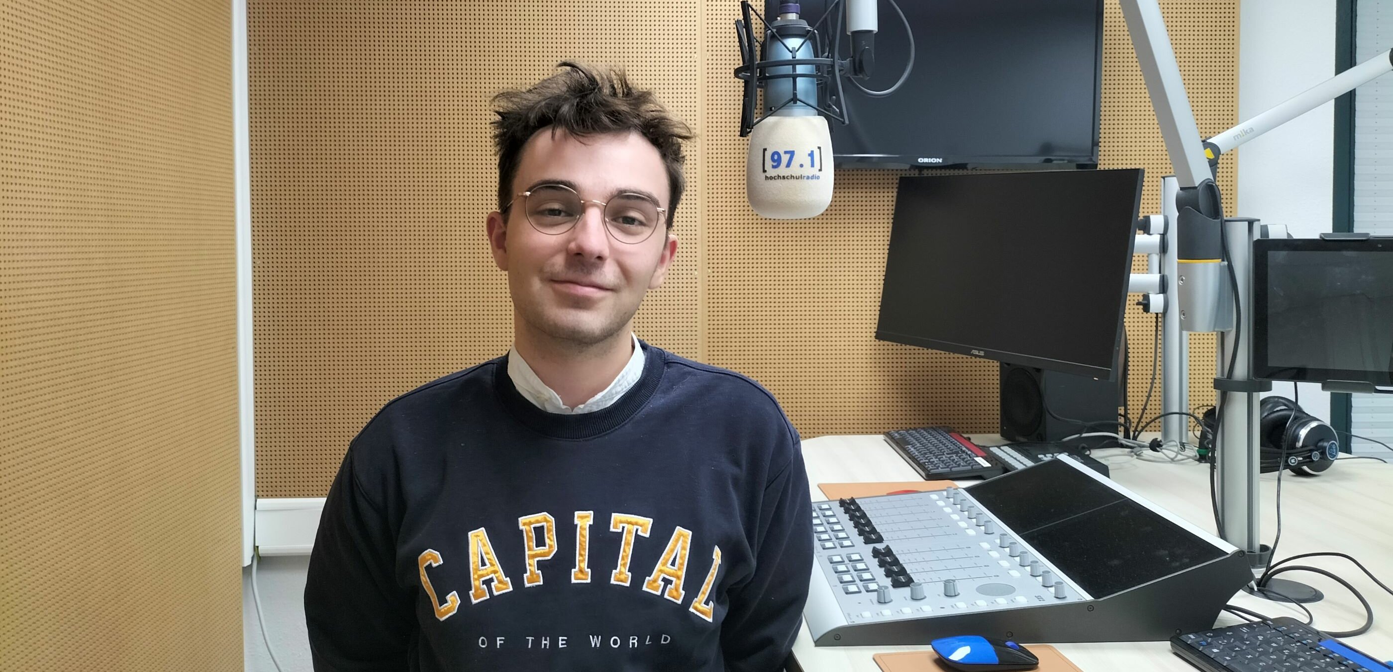 Auf dem Foto ist Josua Wienen zu sehen. Er steht im Studio, neben einem Mikrofon und lächelt in die Kamera.