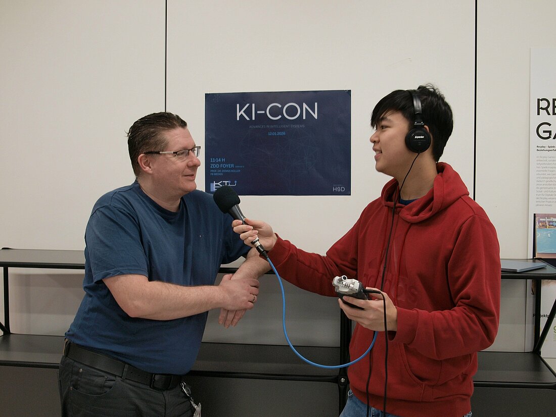 Ein Mann hält einem anderen Mann ein Mikrofon hin und interviewed diesen vor einer weißen Wand mit einem blauen Plakat auf dem KI-CON 2026 steht.