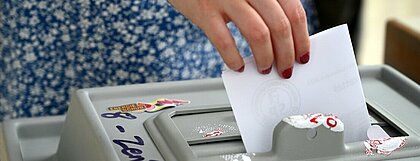 Auf dem Foto ist eine Hand zu sehen, die einen Wahlzettel in eine Urne steckt. 