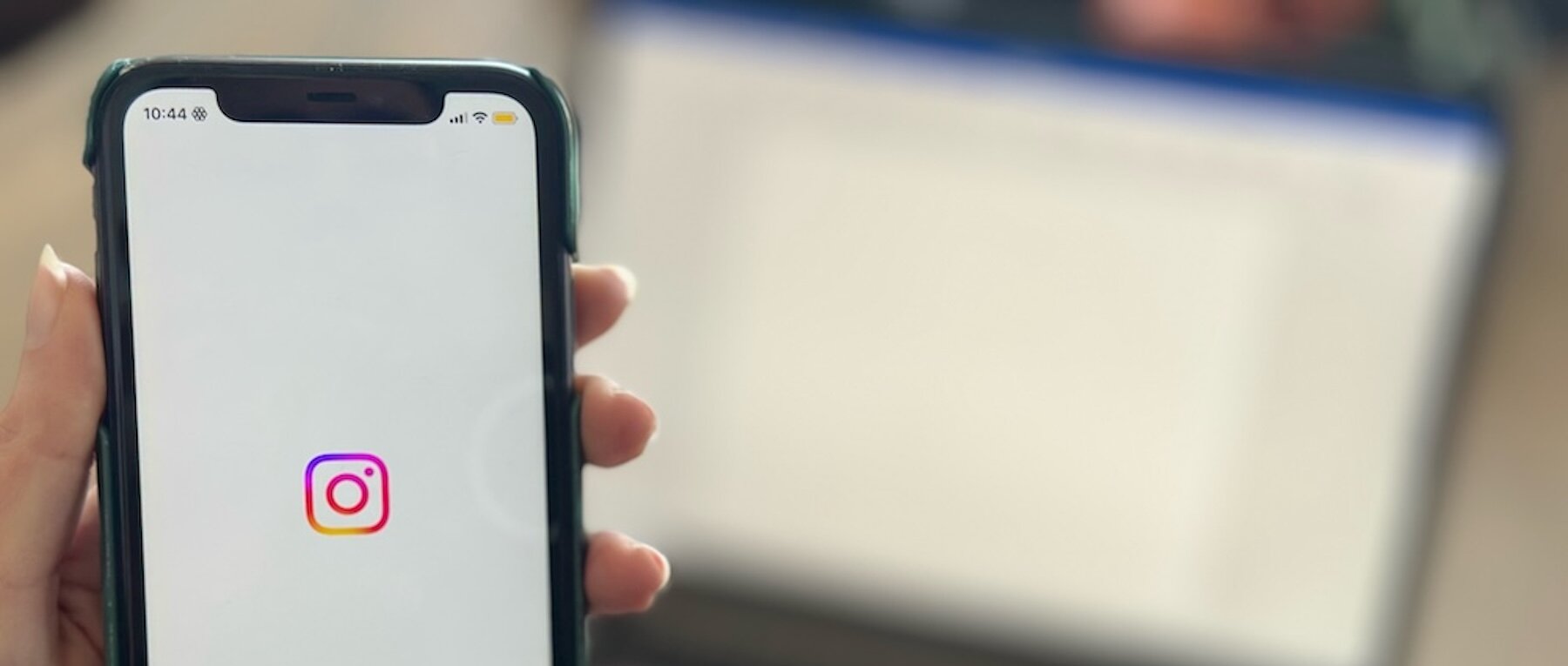 Man sieht eine Hand, die ein Smartphone hält, auf dem das Instagram Logo abgebildet ist. Im Hintergrund ist ein verschwommener Laptop-Bildschirm zu sehen.