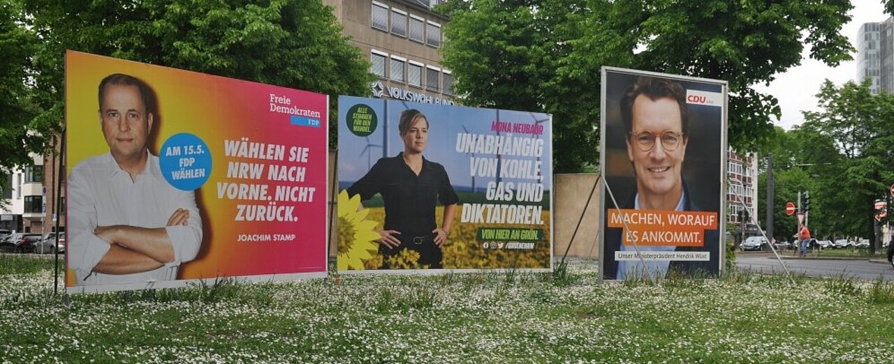 Die Spitzenkandidat:innen präsentieren sich auf großen Wahlplakaten. (Foto: Andreas Meske) Zu sehen sind drei große Wahlplakate auf einer Wiese