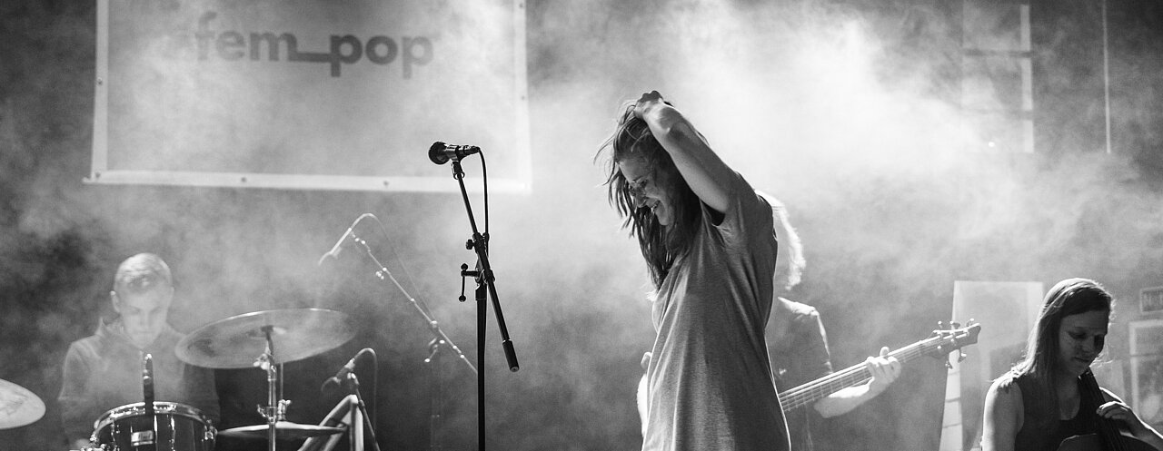 Zu sehen ist die Band bei einem Konzert von fem_pop im zakk 2020. Juliane Blum auf einer schwarz-weiß Fotografie, im Hintergrund Hans Fahr am Schlagzeug, Nils Schröder am Bass und Kamilla Eggeling am Cello und nicht zu sehen Lukas Lohner am Piano.