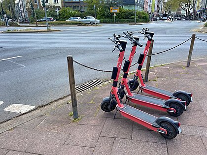 Geparkte E-Scooter auf dem Bürgersteig. Zu sehen sind drei E-Scooter geparkt auf dem Bürgersteig an einer Hauptstraße.