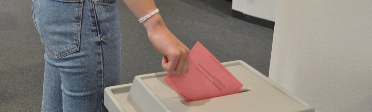 Auf dem Foto ist eine Hand zu sehen, die einene roten Wahlzettel in eine Urne wirft. 