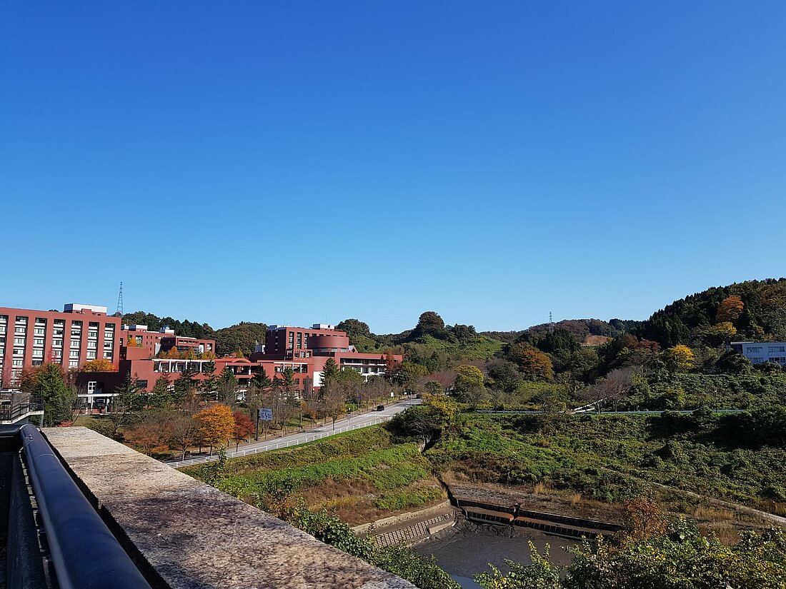 Man sieht eine weitere Aufnahme der roten Gebäude der Kanazawa-Universität. Auf der rechten Hälfte des Bildes erkennt man eine größere, grüne Hügellandschaft.