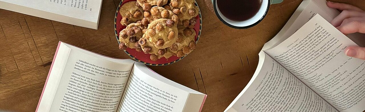 Ein Tisch gedeckt mit Keksen und Tee. Vier Bücher liegen aufgeschlagen auf dem Tisch. Eine Person blättert in einem dieser Bücher. 