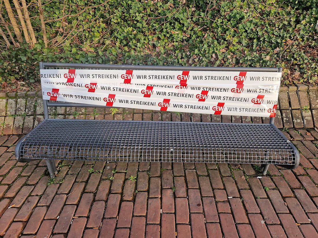 Zu sehen ist eine Sitzbank auf dem Campus. Um die Bank herum wurde ein Band von der GEW gebunden. 