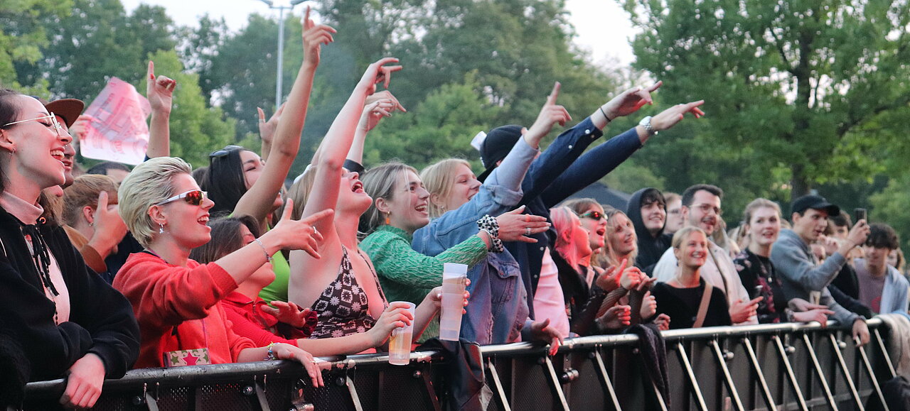 Festivalstimmung auf dem Sommerkult 2022. (Foto: Lena Berghaus) Zu sehen sind junge Menschen, die vor einer Bühnenabsperrung stehen und singen und tanzen
