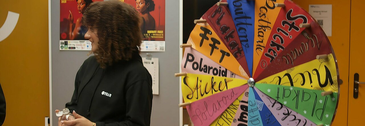 Auf dem Bild sieht man ein Rad mit verschiedenen Kategorien, welches man drehen kann um Gewinne zu kriegen. Davor steht eine Frau, im Hintergrund hängt ein Plakat von dem Filmfest