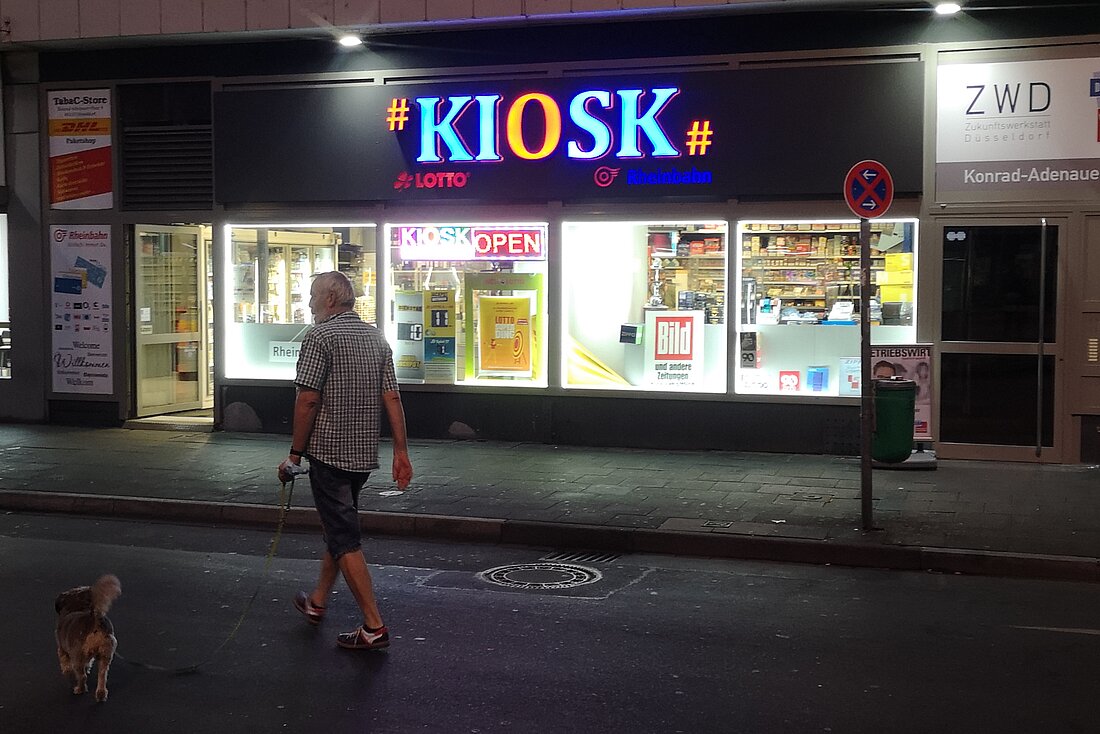 Man sieht einen hell leuchtenden Laden über dem groß „Kiosk“ steht. Ein Mann läuft gerade mit seinem kleinen Hund daran vorbei.