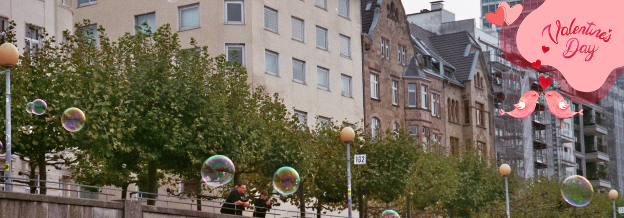 An der Düsseldorfer Rheinpromenade schweben Seifenblasen vor Bäumen und Wohnhäusern. Oben im Bild ist ein „Valentine’s Day“-Schriftzug, darunter zwei küssende Vögel und zwei Herzen.