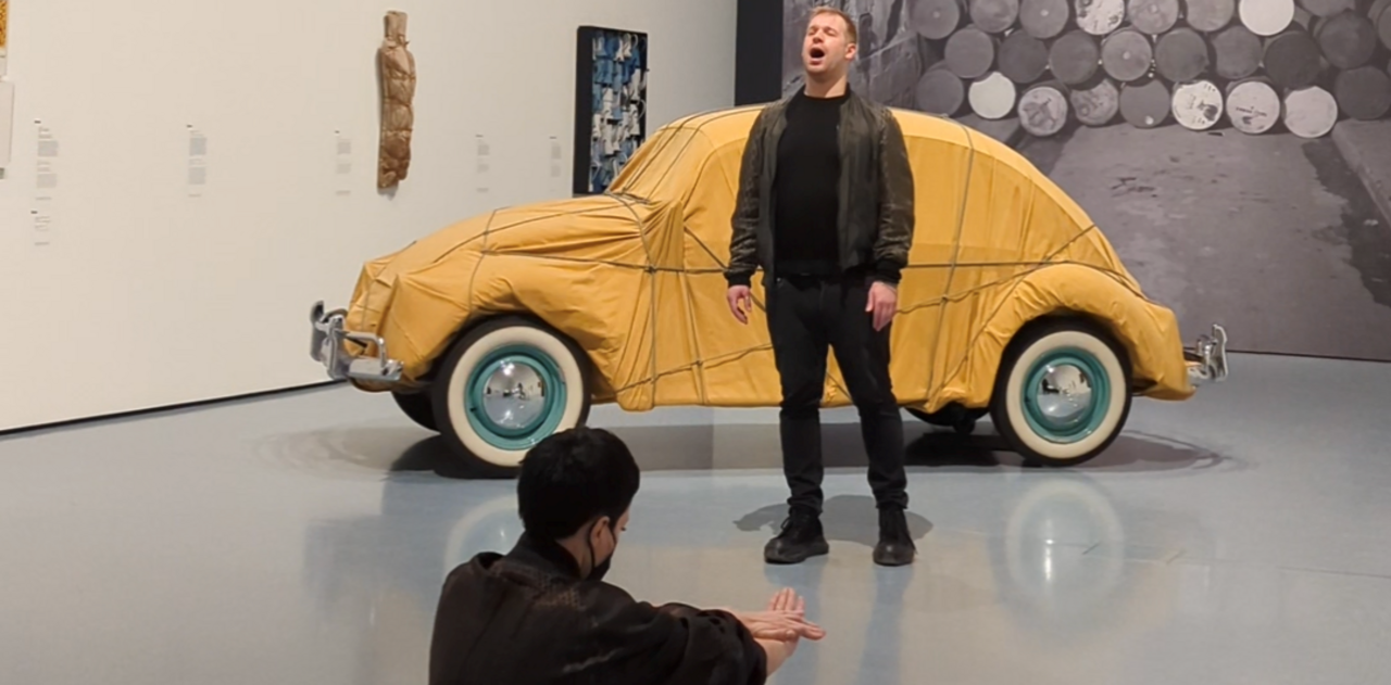 Thomas Huy steht singend vor dem von Christo im Jahr 1961 verhüllten VW-Käfer. Vor Huy bewegt sich die Künstlerin Marlon Red theatralisch auf ihn zu.