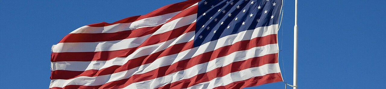 Eine große amerikanische Flagge weht von rechts nach links im Wind. Sie besteht aus roten und weißen Streifen sowie einem dunkelblauen Feld mit vielen weißen Sternen. Rechts im Bild befindet sich der hohe weiße Fahnenmast. Der Hintergrund besteht vollständig aus einem kräftig blauen Himmel.