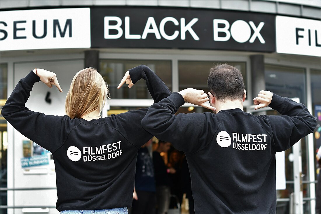 Zu sehen sind ein Junge und ein Mädchen vor der Blackbox des Filmmuseums in Düsseldorf von hinten, die einen Filmfest Pullover anziehen und mit ihren Armen darauf zeigen