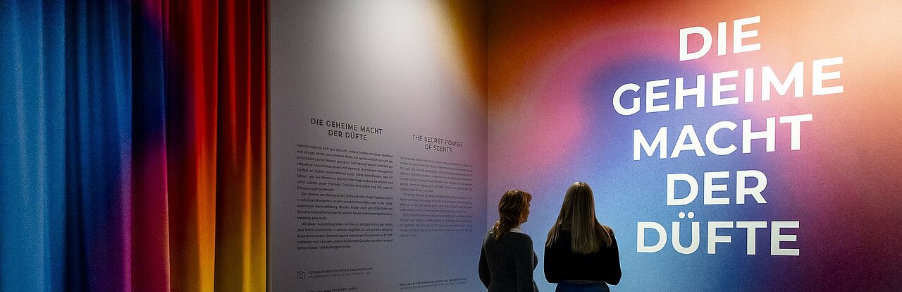 Auf dem Bild sieht man zwei Menschen, die auf eine bunte Wand im Kunstpalast schauen, auf der mit weißer Farbe "Die geheime Macht der Düfte" geschrieben steht.