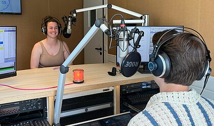 Auf dem Foto ist Geraldine Hupp zu sehen. Sie sitzt im Studio von hochschulradio düsseldorf vor einem Mikrofon und lächelt in die Kamera.Im Vordergrund ist der Kopf des Moderators zu sehen wie er ins Mikro spricht.
