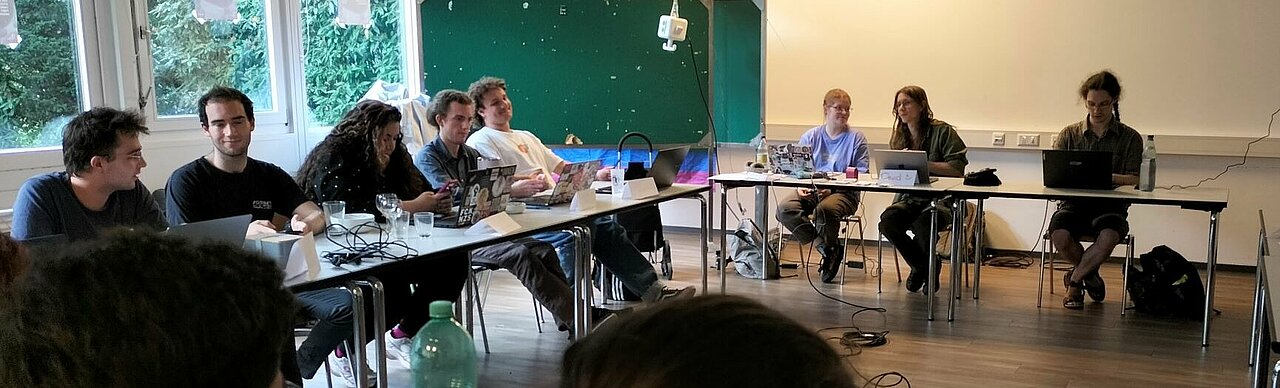 Auf dem Foto sind Mitglieder des Studierendenparlaments zu sehen. Sie sitzen an langen Tischen, die in einem Viereck aufgestellt sind und unterhalten sich.
