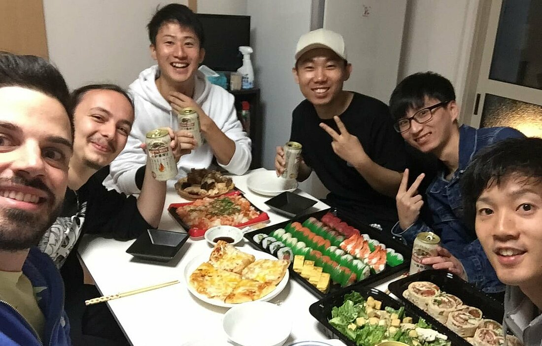 Man sieht Studierende aus verschiedenen Ländern der Welt an einem voll gedeckten Tisch für ein Selfie posieren. Sie halten japanisches Bier in der Hand und auf dem Tisch steht ein Buffet aus Sushi und anderen Speisen.