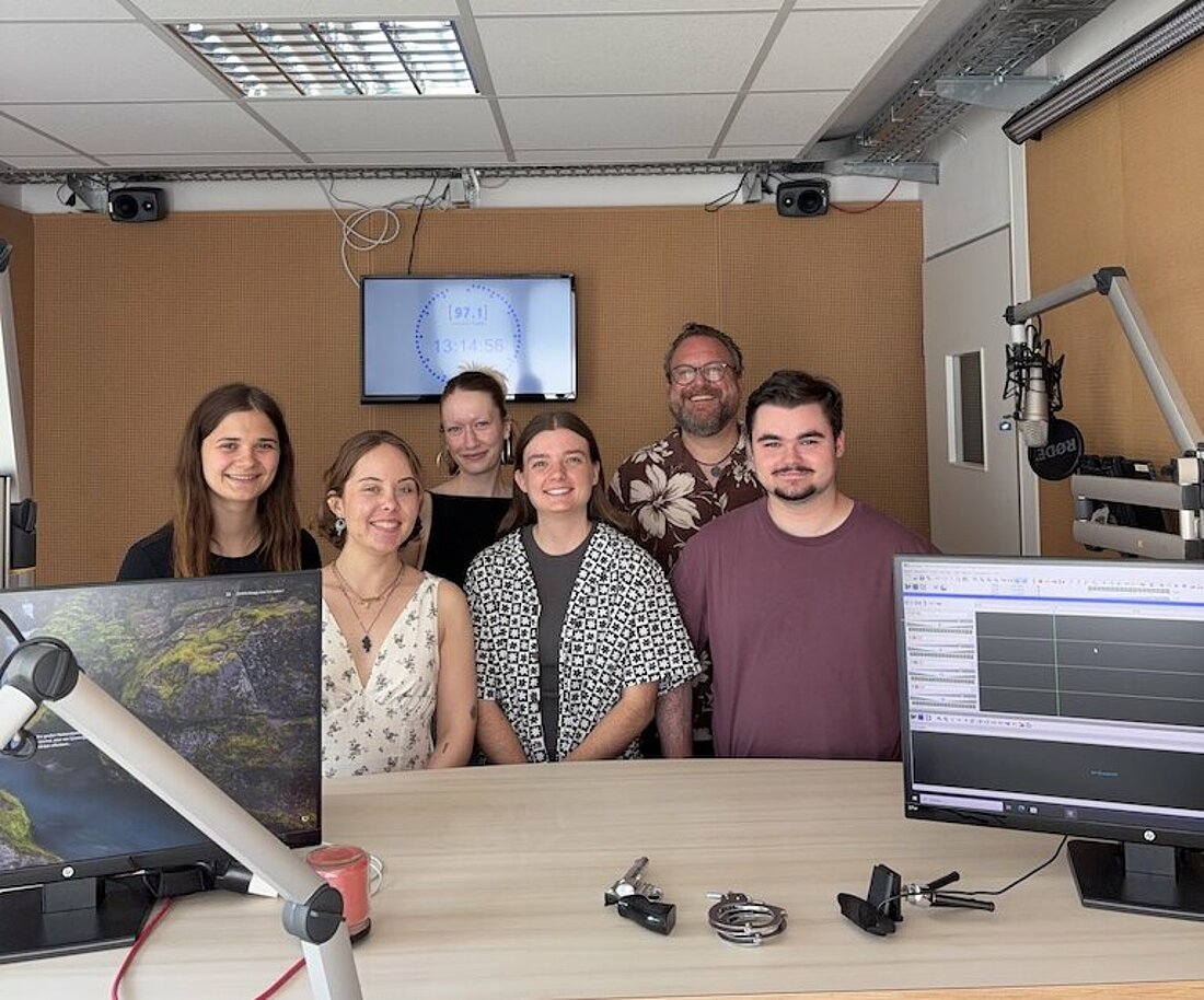 Auf dem Bild sind sechs Personen auf einem Gruppenbild zu sehen. Sie stehen im Tonstudio.