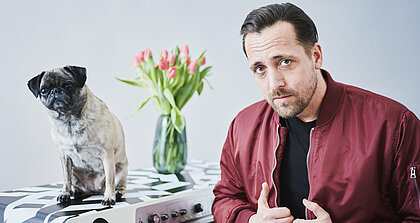 Ein Mann blickt in die Kamera und sitzt in weinroter Jacke vor einem schwarz-weißen Klavier, auf dem eine Vase mit Blumen steht und ein Mopshund sitzt, der ebenfalls in die Kamera schaut.
