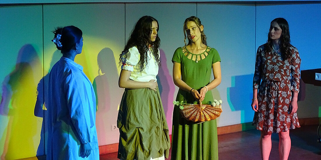 Auf dem Bild sieht man 4 Schauspielerinnen. Der Fokus liegt auf einer, welche mit traurigem Gesichtsausdruck in der Mitte steht. Sie wird besorgt von den anderen angeschaut.