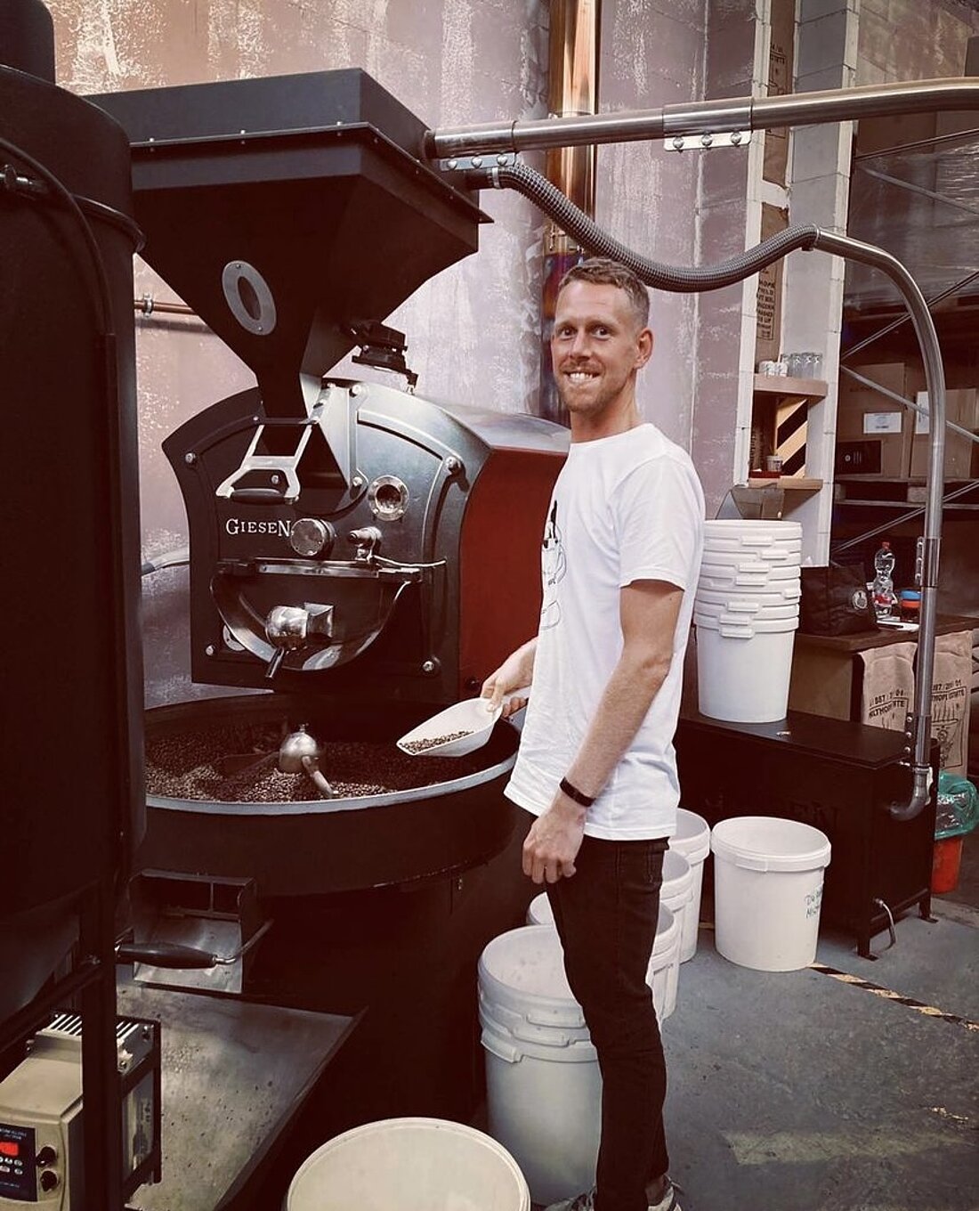 Regelmäßige Kontrollen sorgen für die besondere Note des Kaffees. (Foto: „Die Kaffee“ - Instagram) Auf dem Bild sieht man einen Mitarbeiter, der an der offenen Röstmaschine steht. In der Hand hält er eine Schaufel, in der sich geröstete Bohnen befinden.