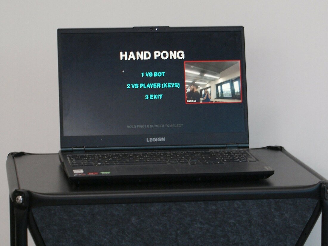 Ein schwarzer Laptop steht auf einem schwarzen Tisch und auf dem Bildschirm sind die Worte "Hand Pong" zu lesen.
