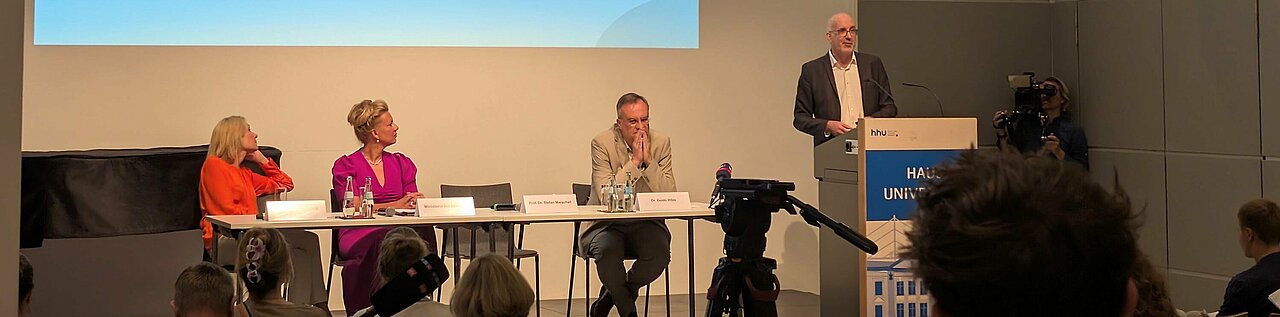 Auf dem Foto sind von links nach rechts die Rektorin der HHU Anja Steinbeck, Wissenschaftsministerin Ina Brandes, der Leiter der Landeszentrale für politische Bildung NRW Dr. Guido Hitze und der Professor Stefan Marschall zu sehen. Stefan Marschall steht am Rednerpult. Die anderen sitzen nebeneinander an einem Tisch.