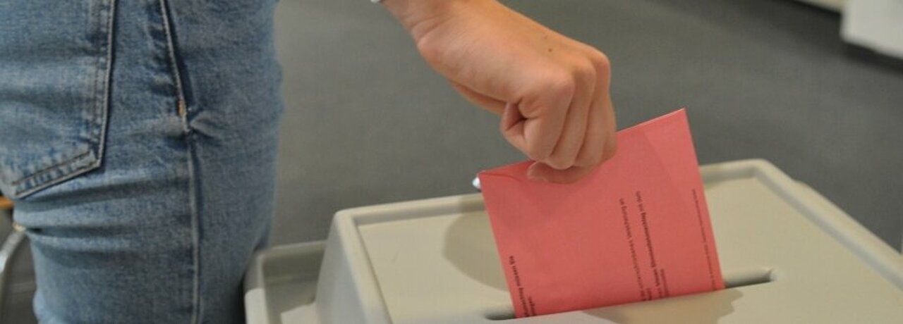 Auf dem Foto ist eine Hand zu sehen, die einen Wahlzettel in eine Wahlurne wirft.