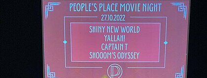Zusehen ist eine Leinwand mit der Projektion: "Plakat der Peoples Place Movie Night" und den Namen der vier FIlme: Shiny New World, Yallah, Captain T, Shrooms Odyssey