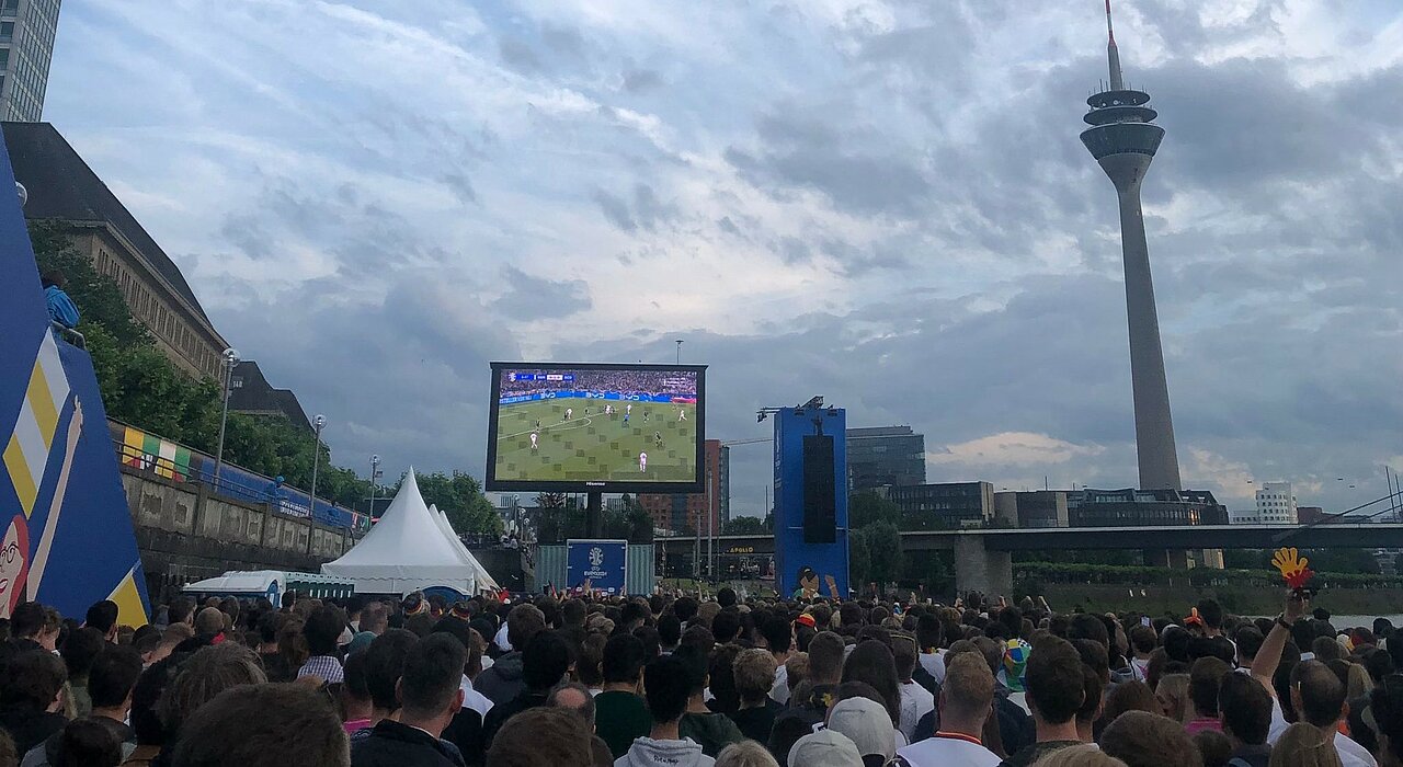 In der Fan Zone am Rheinufer in Düsseldorf. (Foto: Valeska Ridzewski) Auf dem Bild ist die Fan Zone am Rheinufer in Düsseldorf zu sehen. Auf einem großen Bildschirm läuft ein Deutschland Spiel. Es sind viele Leute zusehen, die gekommen sind um das Spiel anzusehen.