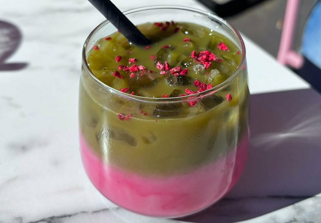 Man sieht ein Glas mit einem Strohhalm auf einem Tisch stehen. Drin befindet sich grüner Matcha Tee der farblich getrennt ist von der rosanen Milch mit Rosensirup. Obendrauf sind ein paar getrocknete, kleingehackte Rosenblätter