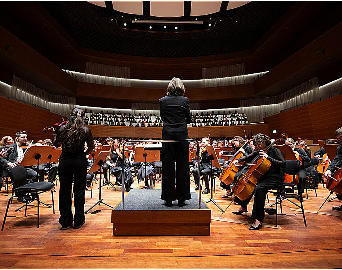 Auf dem Bild sieht man die Dirigentin Silke Löhr von hinten, die auf Chor und Orchester blickt.