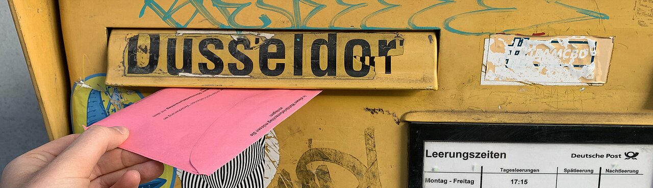 Auf dem Foto ist ein gelber Briefkasten zu sehen. Jemand wirft gerade eine roten Wahlbrief ein.