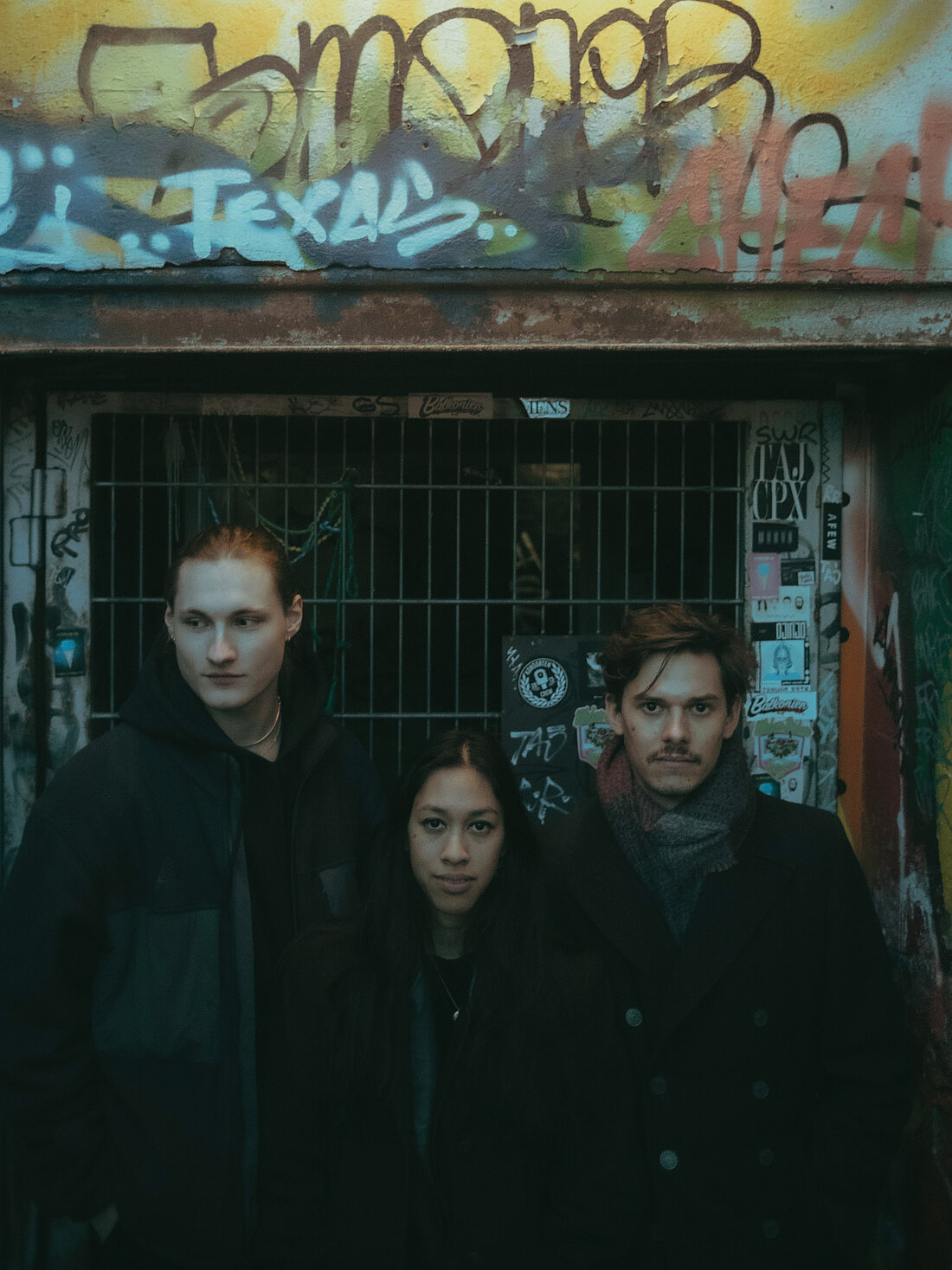 Die drei Bandmitglieder Tim Dierich, Nadia Wardi und Matthias Köhnen sind schwarz gekleidet und stehen vor einem dunklen Hintergrund. An den Wänden befinden sich viele Sticker. 