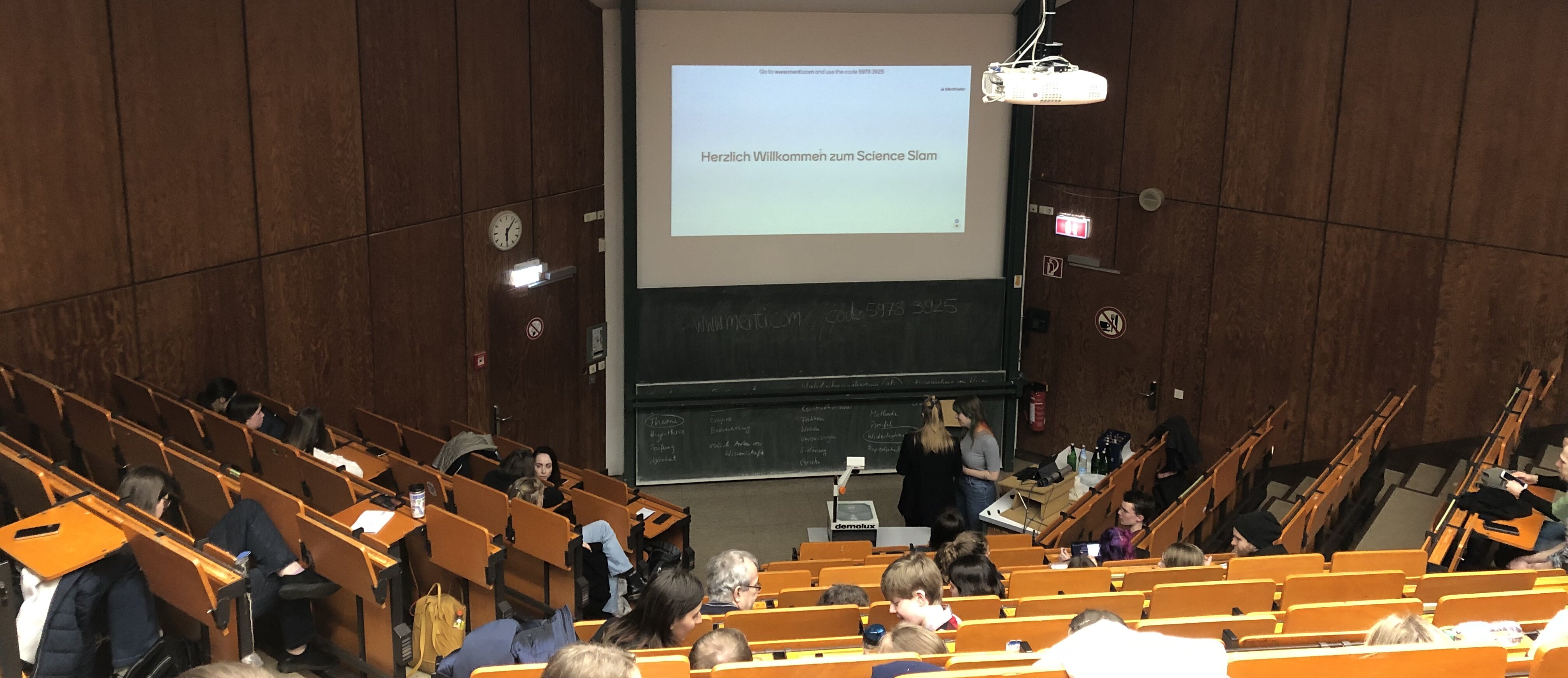 Es nahmen drei Personen am Science Slam teil. (Valeska Ridzewski) Auf der Folie steht "Herzliche Willkommen zum Science Slam"