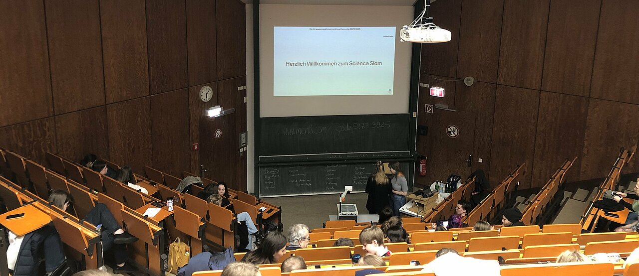 Es nahmen drei Personen am Science Slam teil. (Valeska Ridzewski) Auf der Folie steht "Herzliche Willkommen zum Science Slam"