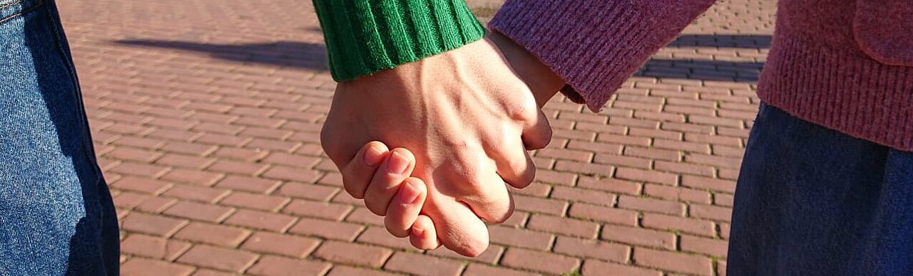 Das Bild zeigt in Nahaufnahme zwei Hände, die sich in der Abendsonne auf dem Campus halten.