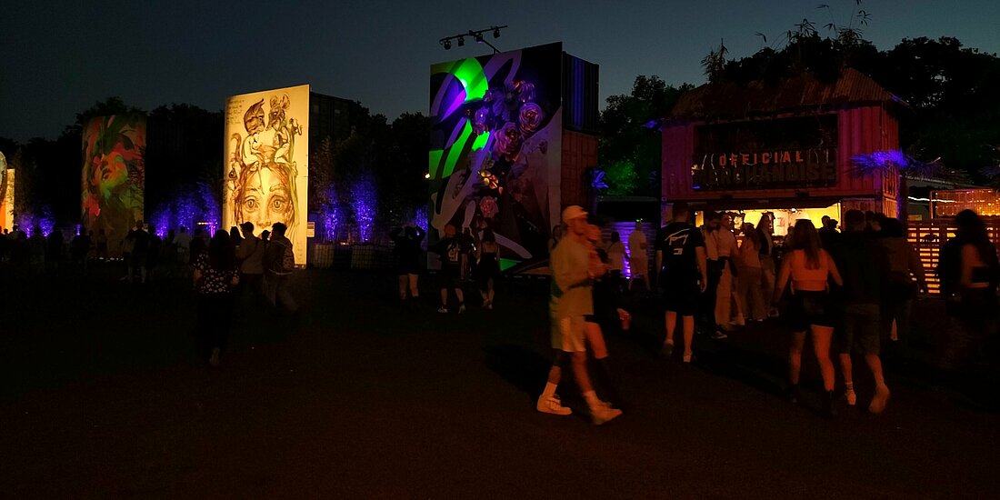 Auf dem Foto ist die Fußgängerzone mit den Leinwänden bei Nacht zu sehen. Einige Besucher:innen laufen umher. Die Leinwände strahlen hell.